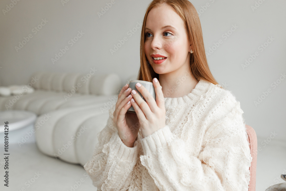 Young charming redhead girl with long hair holding cup of hot drink to warm herself up during cold winter days, sitting in cozy white living-room of her best friend in lovely sweater Young charming redhead girl with long hair holding cup of hot drink to warm herself up during cold winter days, sitting in cozy white living-room of her best friend in lovely sweater