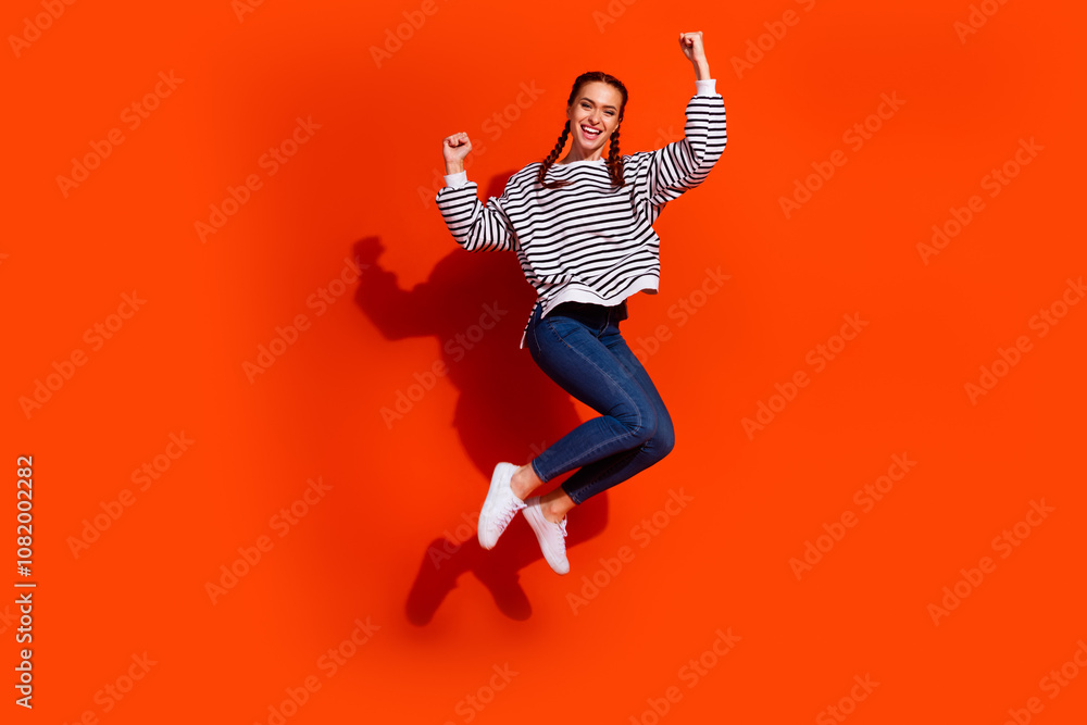 Fototapeta premium Joyful young woman jumping in trendy casual wear against bright orange background, showing happiness and confidence