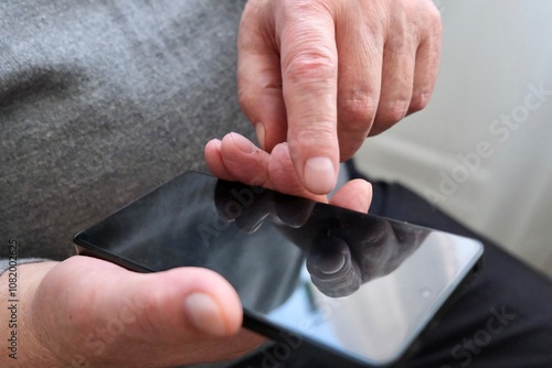 An elderly man is sitting by the window with a smartphone in his hands and talking. The smartphone is in the hands of an elderly retired man.
