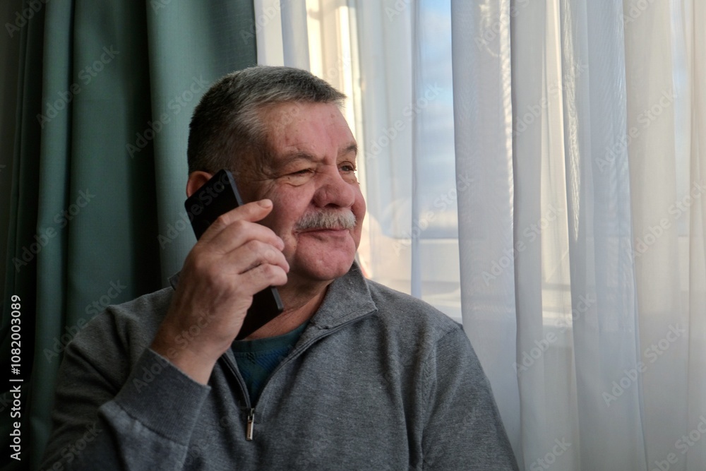 An elderly man is sitting by the window with a smartphone in his hands and talking. The smartphone is in the hands of an elderly retired man.
An elderly man is sitting by the window with a smartphone in his hands and talking. The smartphone is in the hands of an elderly retired man.