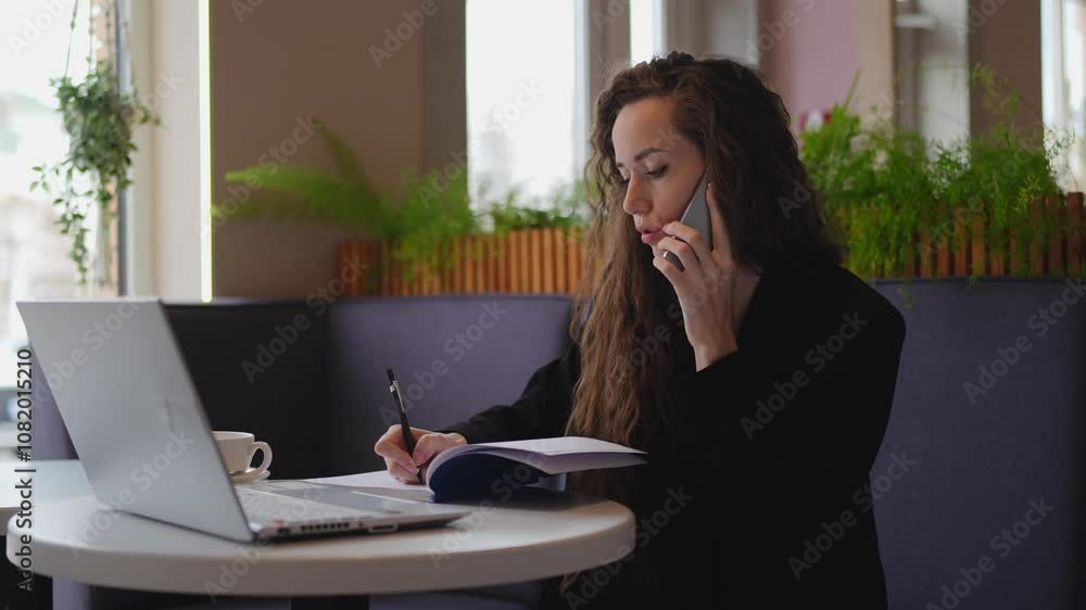 Woman on a phone call while taking notes in a café, illustrating multitasking and professionalism. Woman on a phone call while taking notes in a café, illustrating multitasking and professionalism.