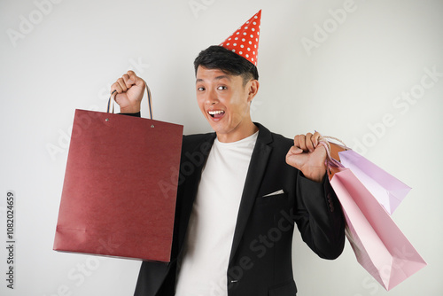 Excited young asian man wearing red party hat, white shirt and black suit is carrying many shopping bags, isolated over white background. Concept for Christmas Holiday and New Year Party