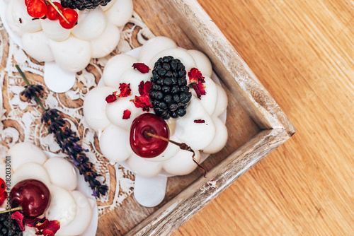 Delicate white mousse dessert topped with fresh blackberries, cherries, and dried rose petals, displayed on a rustic wooden tray with a lace mat. A lavender sprig adds an aromatic touch.