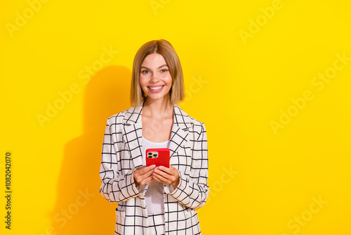 Charming young businesswoman holding smartphone, wearing stylish white jacket, smiling against bright yellow background