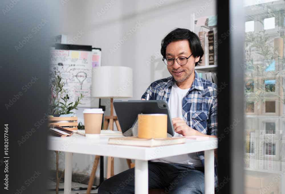 One Asian Japanese male startup worker concentrates on working with digital tablet at desk near daylight window in casual small business office, online freelance jobs, and E-commerce SME entrepreneur. One Asian Japanese male startup worker concentrates on working with digital tablet at desk near daylight window in casual small business office, online freelance jobs, and E-commerce SME entrepreneur.