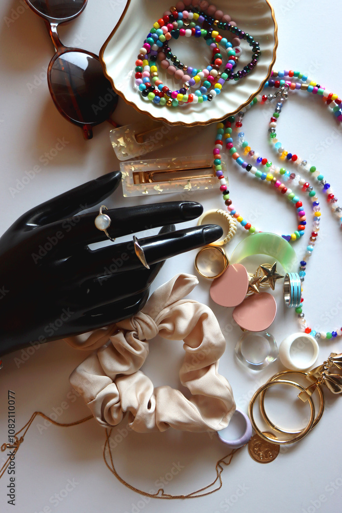 Hand figurine, sunglasses, gold jewelry, colorful bead jewelry, statement rings and various hair accessories on the white table. Flat lay.