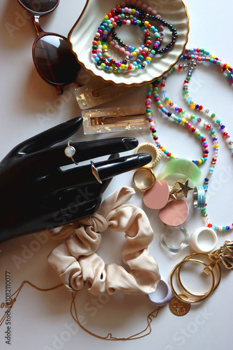 Hand figurine, sunglasses, gold jewelry, colorful bead jewelry, statement rings and various hair accessories on the white table. Flat lay.