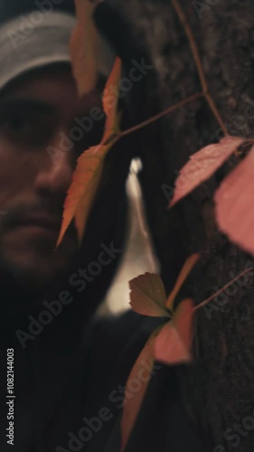 Eerie Autumn – Young man in black hoodie and gray hat in autumn forest, close-up near pine tree, red leaves sliding down trunk hiding behind leaves in shadow, eerily swaying leaves in the wind 