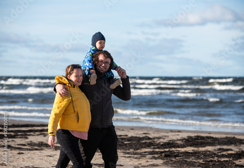 A happy family strolls along a chilly autumn beach, with their young son joyfully running ahead, creating lasting memories as the crisp wind and soothing waves embrace them in a moment of pure photo