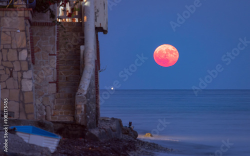 Super Moon rise above a tranquil coast, where a small boat drifts in the calm waters, surrounded by a dark blue sky with clouds and distant lights. photo