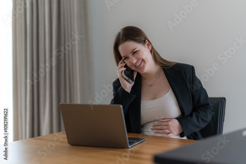 Cheerful gen z pregnant woman working on laptop at home and talking to the phone. 