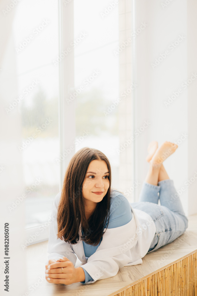 girl lying on her stomach on the windowsill, pensive look, slight smile in a white shirt and light jeans girl lying on her stomach on the windowsill, pensive look, slight smile in a white shirt and light jeans