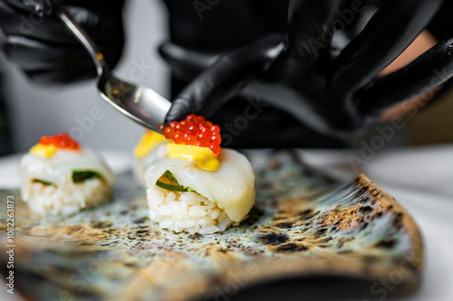 A chef in black gloves artfully garnishes sushi with vibrant yellow sauce and red caviar on a decorative plate, showcasing a meticulous approach to culinary presentation. photo