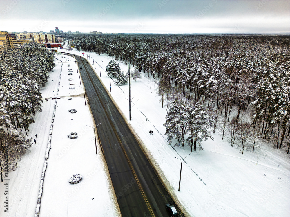 Obraz premium Snow-covered road along the forest with pedestrians walking in winter landscape near a city during a cloudy day