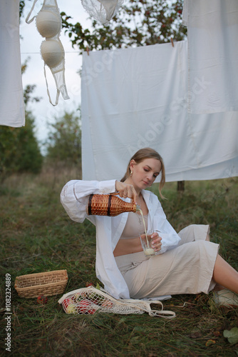 pregnant woman sitting on the grass in the garden and eating an apple and pouring milk into a glass. woman sitting in the garden on the background of laundry drying after washing. hanging laundry afte