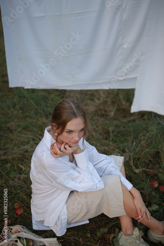 pregnant woman sitting on the grass in the garden and eating an apple and pouring milk into a glass. woman sitting in the garden on the background of laundry drying after washing. hanging laundry afte