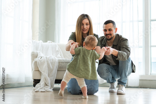 Baby development. Little infant girl making first steps at home. Happy mother father looking at toddler child walking in living room. Happy family at home. Mom dad supporting baby first steps photo