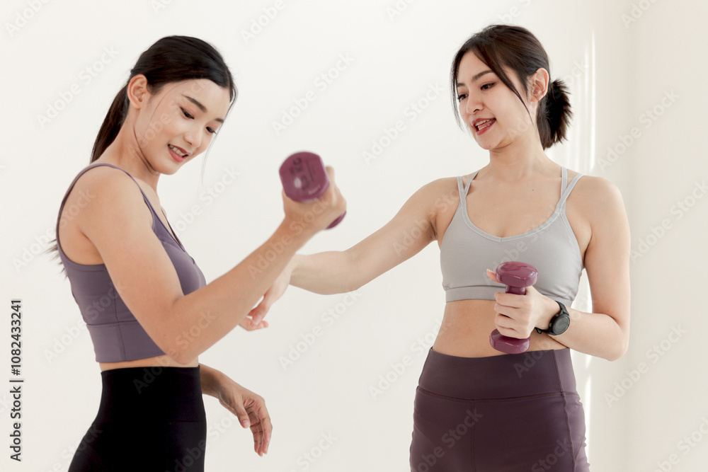 Two Asian women buddy friend exercise with dumbbells together in white room. Happy sporty girl lifting up dumbbell for work out under guidance of trainer on white wall. Healthy sport hobby lifestyle. Two Asian women buddy friend exercise with dumbbells together in white room. Happy sporty girl lifting up dumbbell for work out under guidance of trainer on white wall. Healthy sport hobby lifestyle.