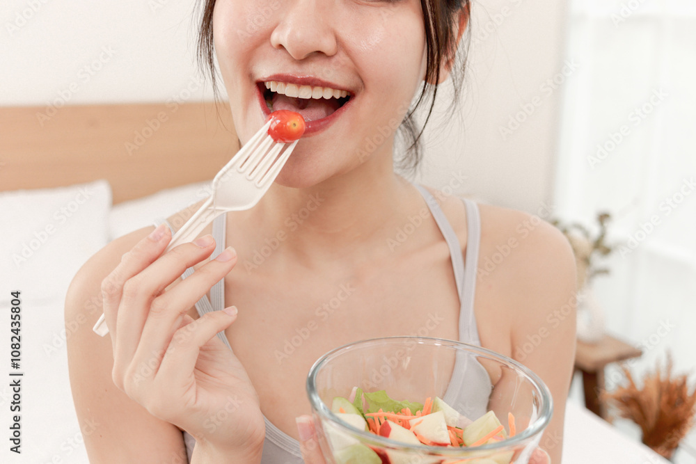 Close up mouth of Asian girl eating salad of fresh fruits and vegetables while sitting on bed in bedroom. Happy beautiful woman holds fork and salad bowl for eating at home. Healthy food and lifestyle Close up mouth of Asian girl eating salad of fresh fruits and vegetables while sitting on bed in bedroom. Happy beautiful woman holds fork and salad bowl for eating at home. Healthy food and lifestyle