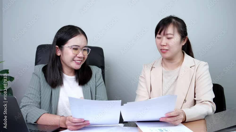 Two Asian businesswomen are working together in an office. One is writing in a notebook while the other reviews documents. A laptop and a binder are also on the desk. Two Asian businesswomen are working together in an office. One is writing in a notebook while the other reviews documents. A laptop and a binder are also on the desk.