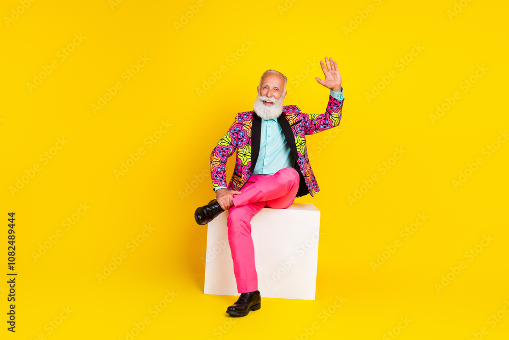 Full body photo of senior male sit white platform wave hand hi wear trendy colorful pink fancy outfit isolated on yellow color background Full body photo of senior male sit white platform wave hand hi wear trendy colorful pink fancy outfit isolated on yellow color background