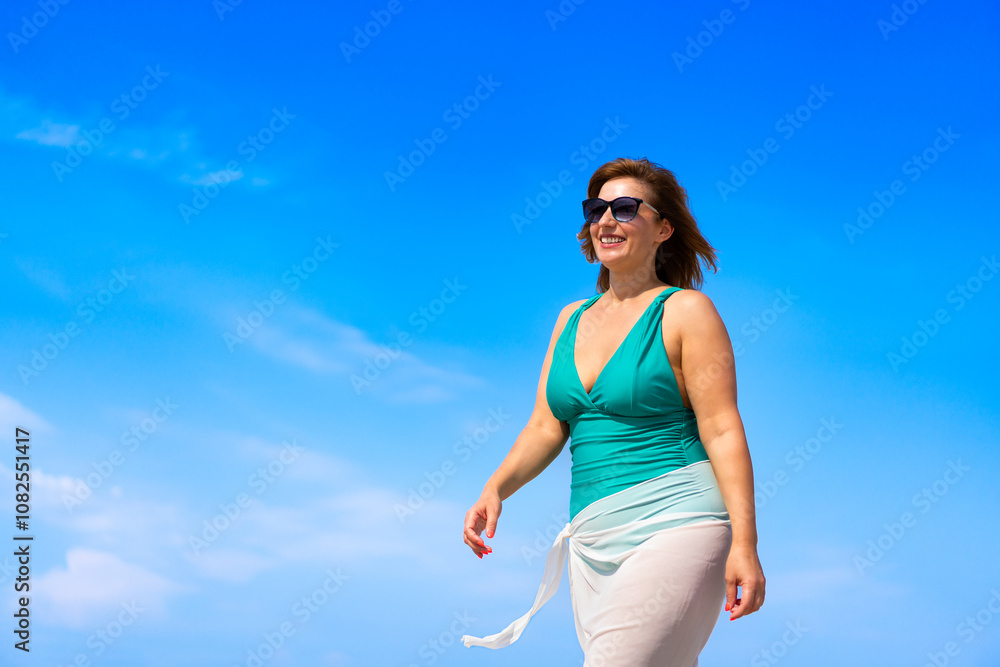 Summer vacation. Portrait of beautiful young woman in one-piece swimsuit and white sarong and sunglasses walking against background of blue sky on sunny day. Sunbathing. Side view. Summer vacation. Portrait of beautiful young woman in one-piece swimsuit and white sarong and sunglasses walking against background of blue sky on sunny day. Sunbathing. Side view.