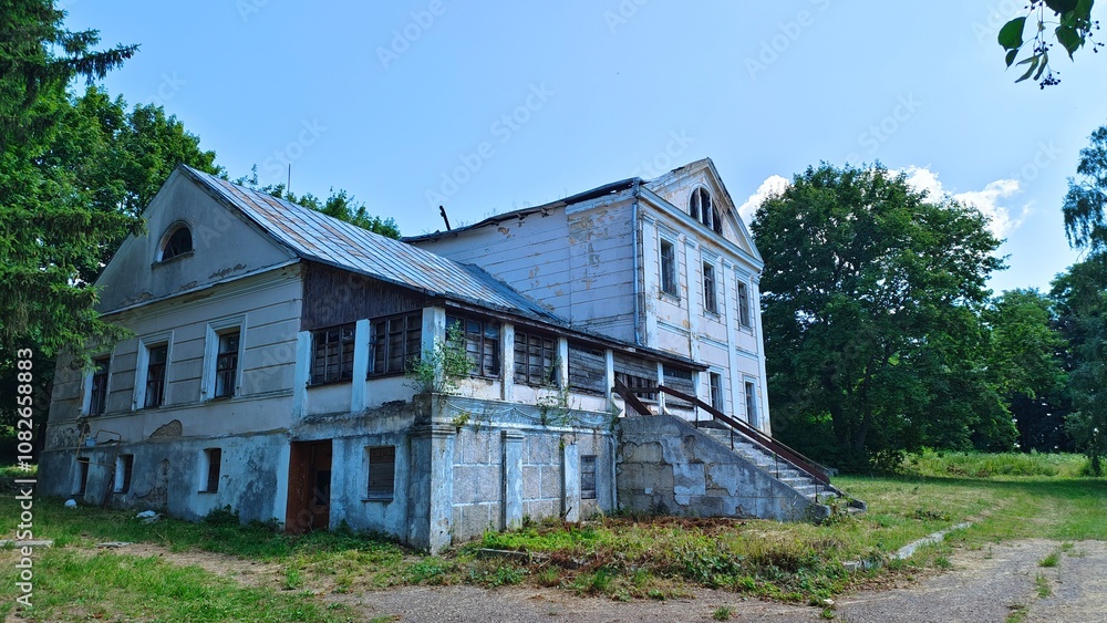 An old abandoned stone house with a metal roof and window frames without glass is surrounded by a grassy lawn and trees. A paved road approaches the front porch with a railing. Sunny summer weather An old abandoned stone house with a metal roof and window frames without glass is surrounded by a grassy lawn and trees. A paved road approaches the front porch with a railing. Sunny summer weather