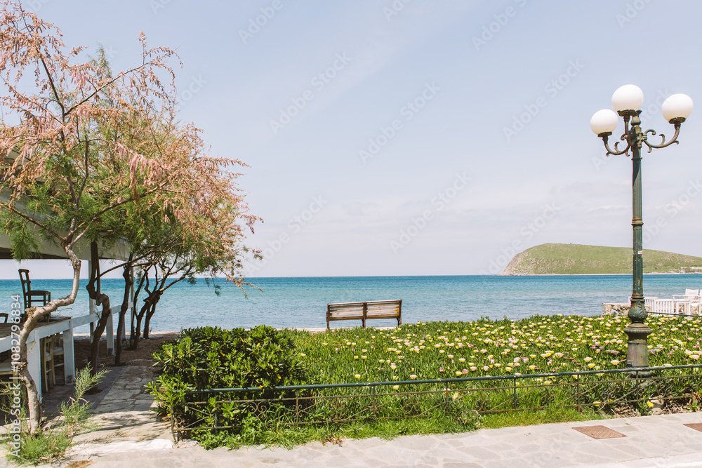 Photo of an empty bench overlooking a beach and clear blue ocean at Limnos, Mirina (marina) Παραλία Ρωμέικος Γιαλός - Scenic Beauty of Lemnos Island, Greece - Aegean Seaside Views