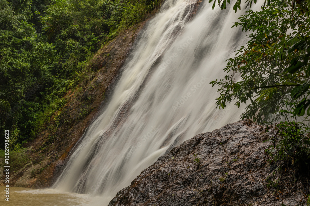 A powerful waterfall tumbling down from a steep rocky cliff surrounded by green foliage. The water falls down with force, creating fog and a sense of movement. A powerful waterfall tumbling down from a steep rocky cliff surrounded by green foliage. The water falls down with force, creating fog and a sense of movement.