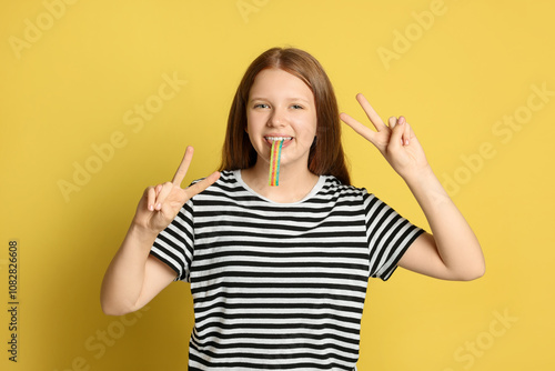 Teenage girl eating tasty rainbow sour belt while showing v-sign on yellow background