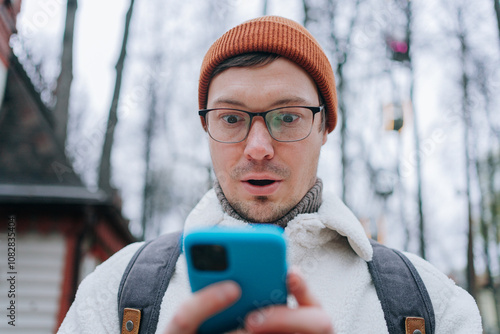 Shocked male tourist wearing orange beanie and eyeglasses reading unbelievable news on smartphone while standing in winter park, expressing astonishment and surprise