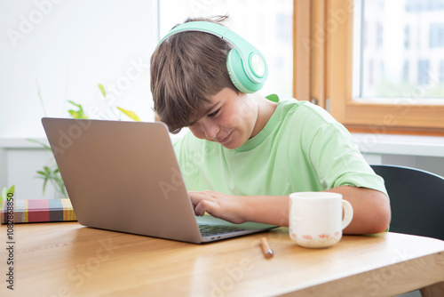 Cute boy in headphones sits at the table and studies online 