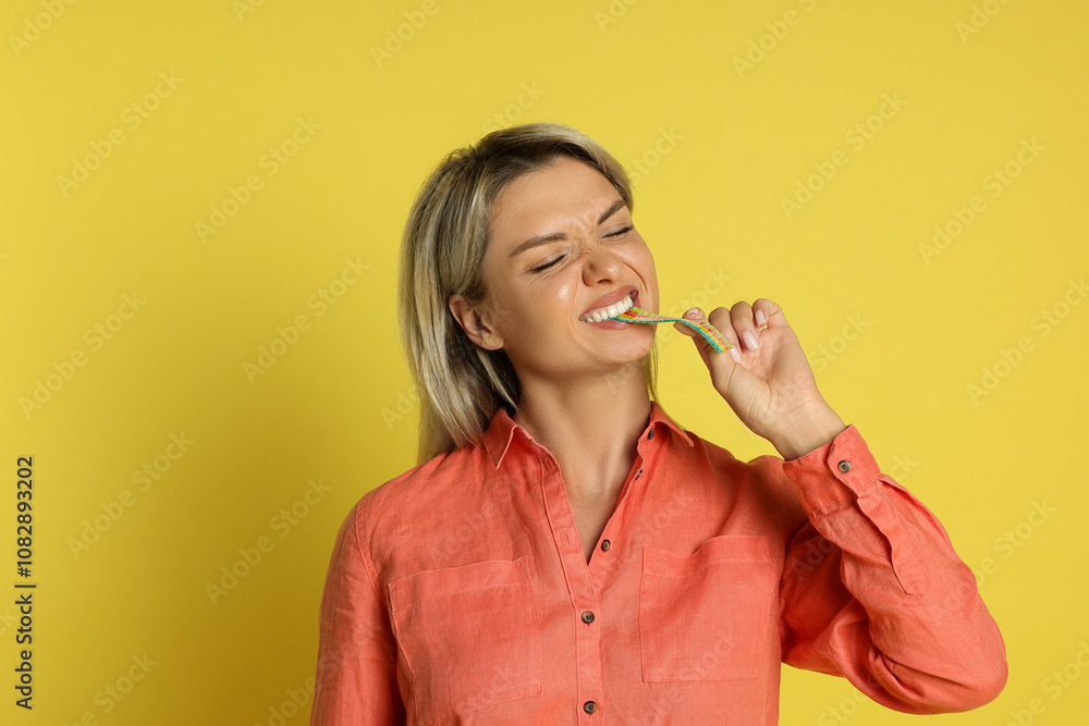 Young woman eating tasty rainbow sour belt on yellow background Young woman eating tasty rainbow sour belt on yellow background