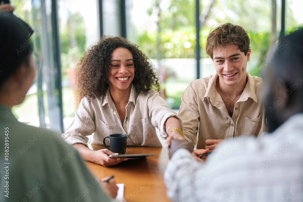 Two young adults, one female with curly hair and one male, engage in a friendly handshake while seated in a sunlit modern office, marking the beginning of a collaborative session.