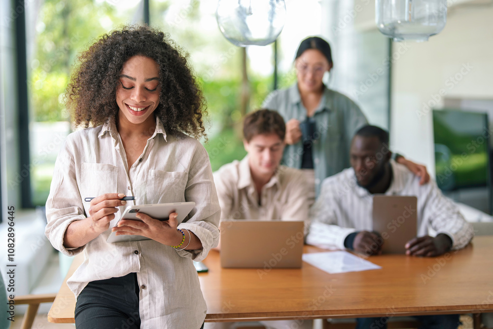 A cheerful African American young adult businesswoman interacts with a digital tablet. Colleagues work in the background of the well-lit, contemporary office space. A cheerful African American young adult businesswoman interacts with a digital tablet. Colleagues work in the background of the well-lit, contemporary office space.