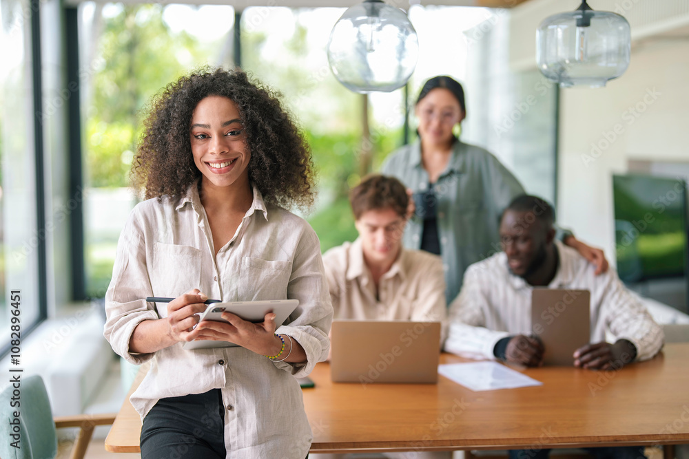 A lively African American woman engages with a digital tablet, her cheerful demeanor evident. Colleagues work behind her in a well-lit office, suggesting a collaborative professional environment. A lively African American woman engages with a digital tablet, her cheerful demeanor evident. Colleagues work behind her in a well-lit office, suggesting a collaborative professional environment.