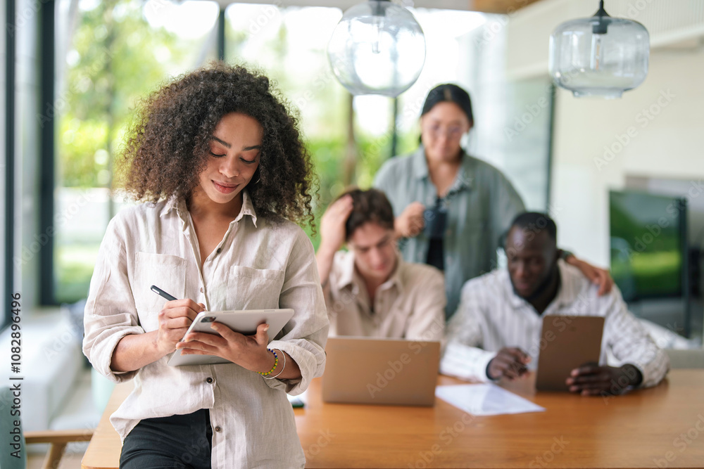 African American young adult businesswoman appears engaged and joyful using a digital tablet. She's in a modern office, dressed smartly with diverse colleagues in the background. African American young adult businesswoman appears engaged and joyful using a digital tablet. She's in a modern office, dressed smartly with diverse colleagues in the background.