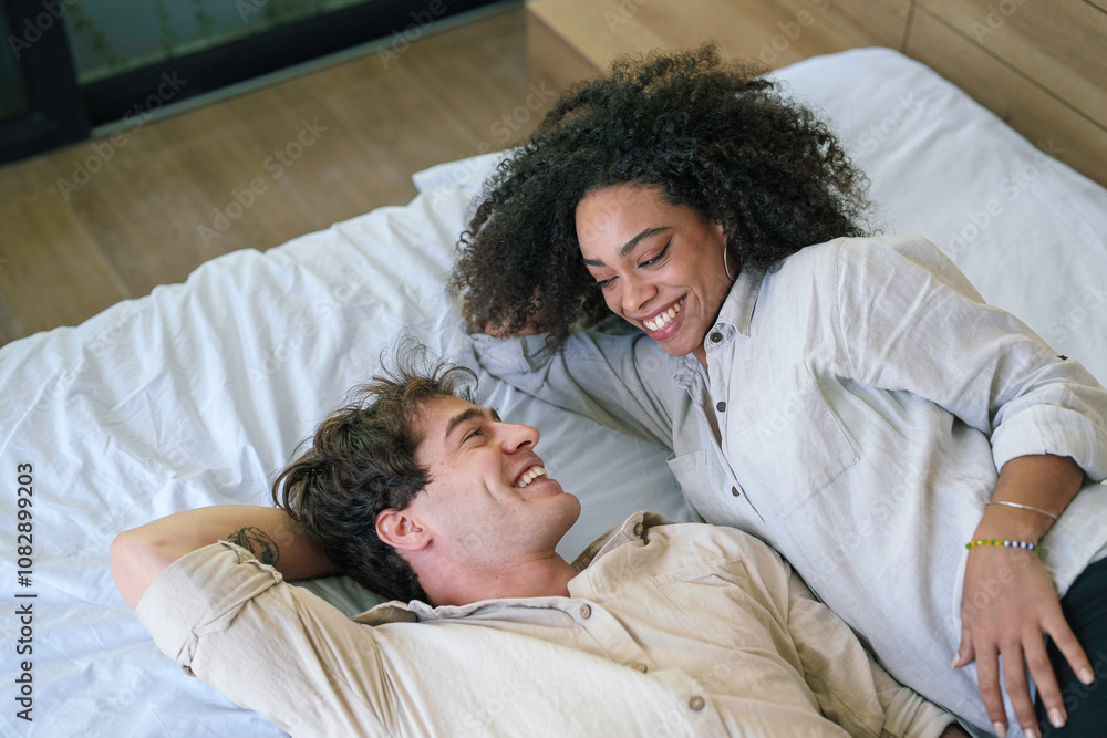 A joyful African American woman and her Caucasian partner relax on a bed, sharing smiles and an intimate moment, dressed casually in light attire. A joyful African American woman and her Caucasian partner relax on a bed, sharing smiles and an intimate moment, dressed casually in light attire.