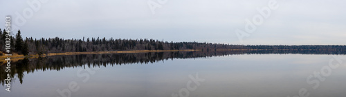 Wide panorama of a calm northern lake reflecting an evergreen forest and autumn colors 