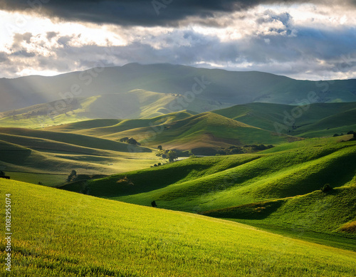 Sunlight breaks through dramatic clouds to illuminate rolling green hills creating a stunning pastoral landscape with patches of light and shadow.