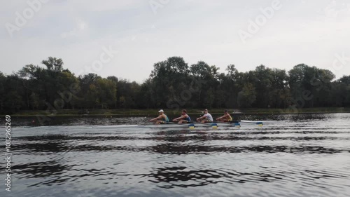 Rowing team training. Side view of 4 young caucasian male rowers, during a rowing practice, athlete sitting in a boat in the river Dnipro, rows through a calm water in autumn. 4k footage. City area in