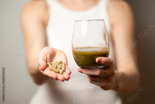 Kratom consumption concept:  female holds a ready to drink glass of green liquid, powdered kratom melted in hot water, glass of tea and handful of pills in the jar, close up photo, selective focus