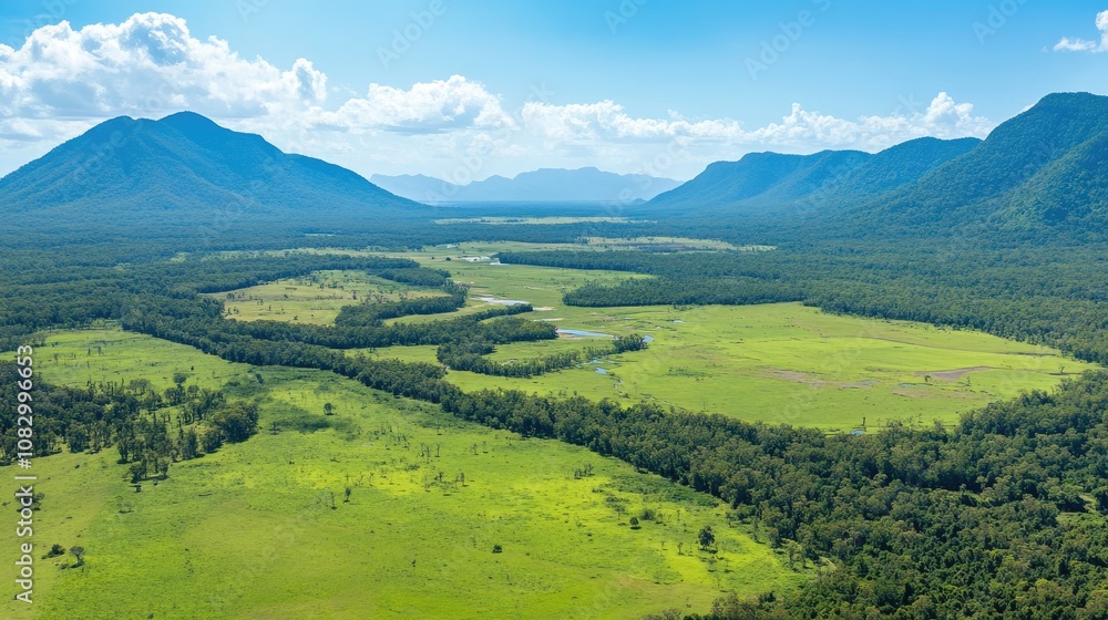 Obraz premium Breathtaking Aerial View of Lush Green Valley Surrounded by Majestic Mountains Under a Bright Blue Sky with Soft Clouds and Scenic River Flowing Through Natural Landscape