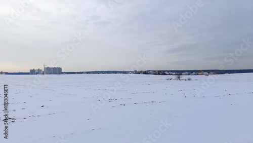 After a snowfall, snow lies on the ground of an agricultural field with buildings and forest behind it. A crane can be seen behind the buildings. The winter sun breaks through the solid cloud cover photo