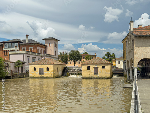 Portogruaor, Venice, Italy - June 26, 2024: Historic yellow stone watermill buildings on Fiume Lemene, greenish river. Waterfall and mansion building on side under blue cloudscape photo