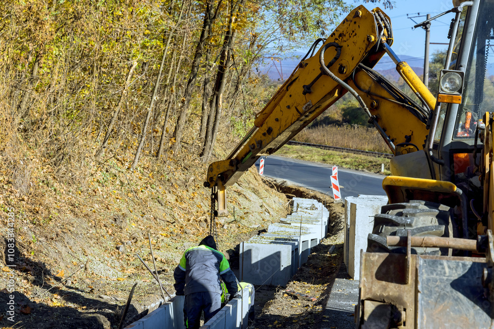 Fototapeta premium Construction worker operates crane to position concrete U blocks beside rural road.