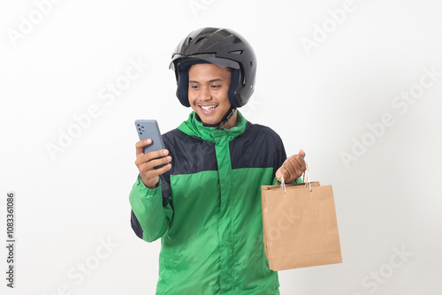 Portrait of Asian online courier driver wearing green jacket and helmet giving craft paper shopping bag while using mobile phone. Isolated image on white background photo