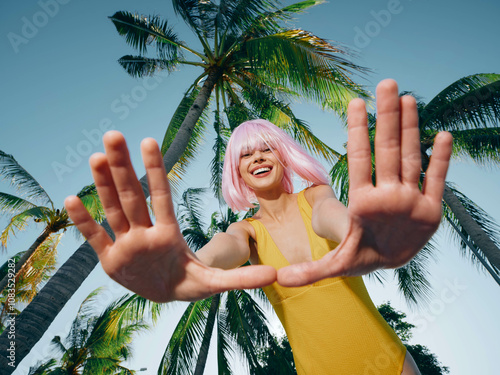 Pinkhaired woman with a joyful smile, standing in front of lush palm trees, raising her hands playfully near her face photo