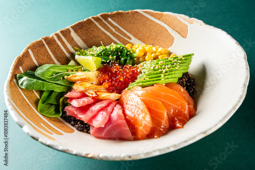 A vibrant poke bowl with salmon, tuna, avocado, corn, seaweed salad, and tobiko on black rice in a ceramic dish against a teal background. Perfect for healthy eating and food photography. photo