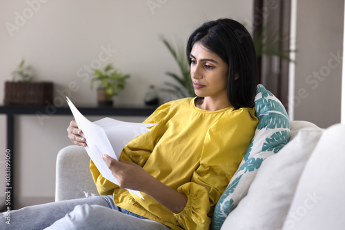 Serious young Indian woman reading legal documents on home couch, reeving loan, insurance agreement, bank notice, paper bill, doing paperwork at home, checking invoices, resting on sofa photo